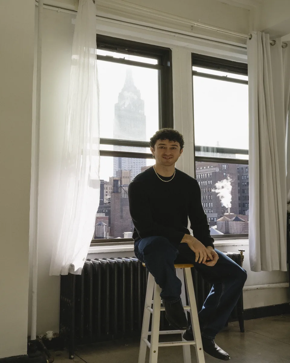 Dylan Kahler on a stool by a studio window, the Empire State Building behind him